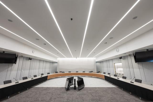 A committee room in the House of Representatives. A long, oval-shaped table with microphones and chairs behind it, is placed along the sides of the room. Four screens are placed on the floor in the middle of the room.