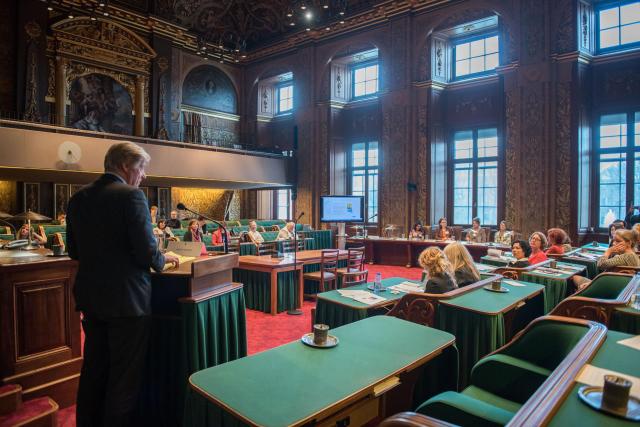 A man is standing in a room full of green desks, some of which members of the Senate are seated at.